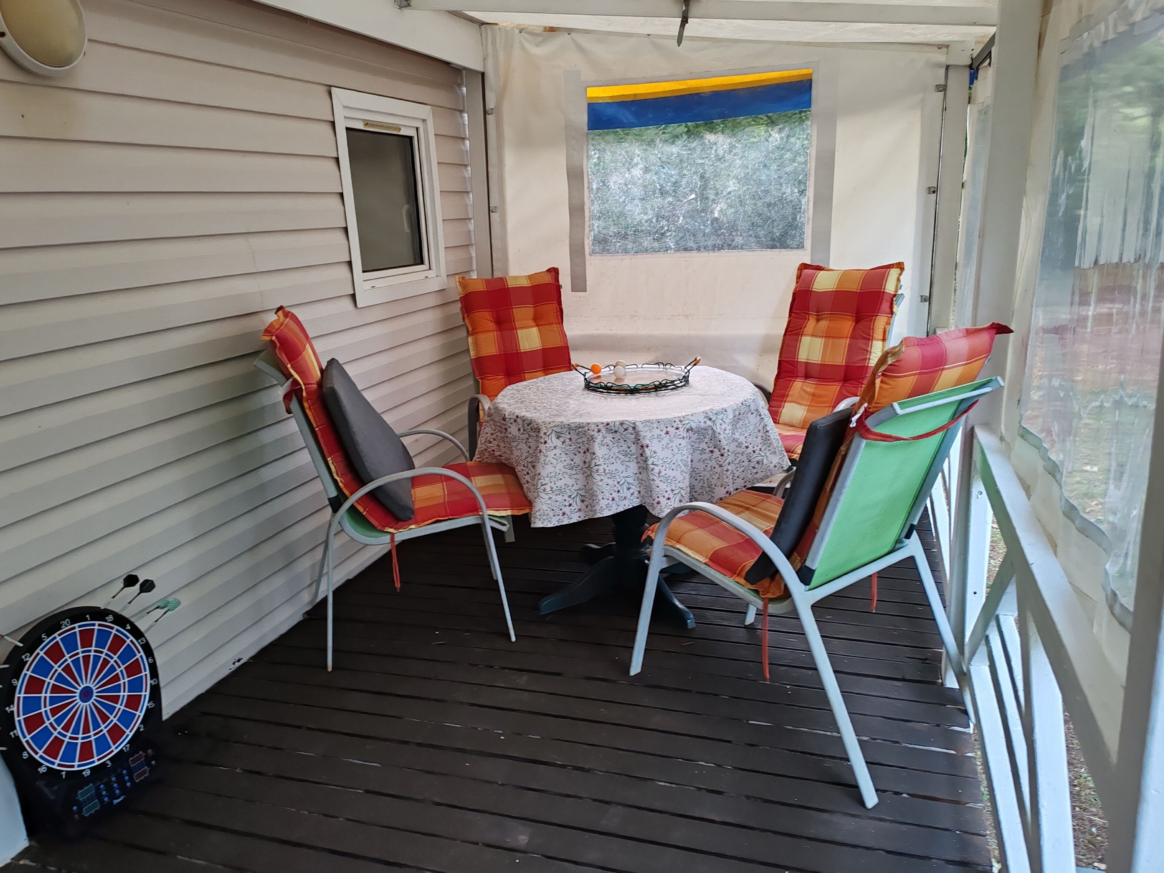 A covered patio with a table and four chairs, a dartboard, and a window with a view of trees.