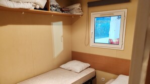 Quarto pequeno com uma cama de solteiro, janela, prateleira sobre a cama e luminária fixada na parede.
