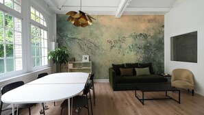 Ein großzügiger Raum mit einem großen weißen Tisch, einem grünen Sofa und einem beige farbenen Sessel. An einer Seite befindet sich ein großes Fenster und ein Wandgemälde.