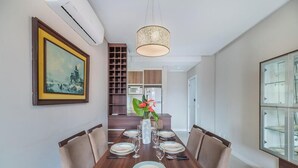 Uma sala de jantar com uma mesa comprida posta para quatro pessoas, um lustre pendurado e um quadro na parede.