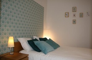 Un lit bien fait, avec des oreillers, une lampe de chevet et un mur recouvert d’un papier peint à motifs et orné de tableaux encadrés.