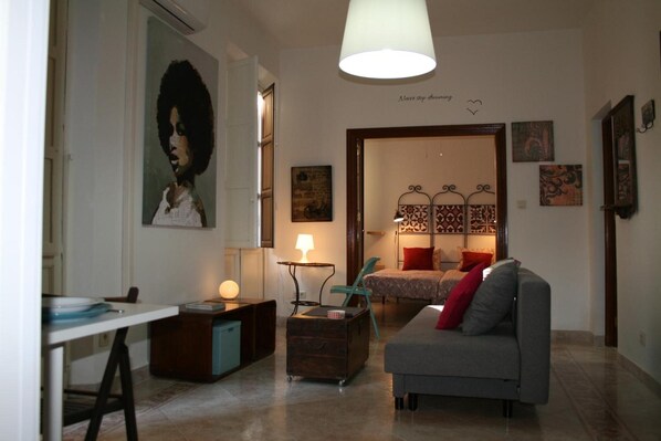 Una sala de estar con sofá, una mesa, una lámpara, una silla y una cama con cortinas estampadas.