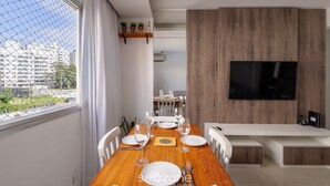 A modern dining area with a wooden table set for six, a TV mounted on a wooden wall, and a view of a cityscape through a large window.
