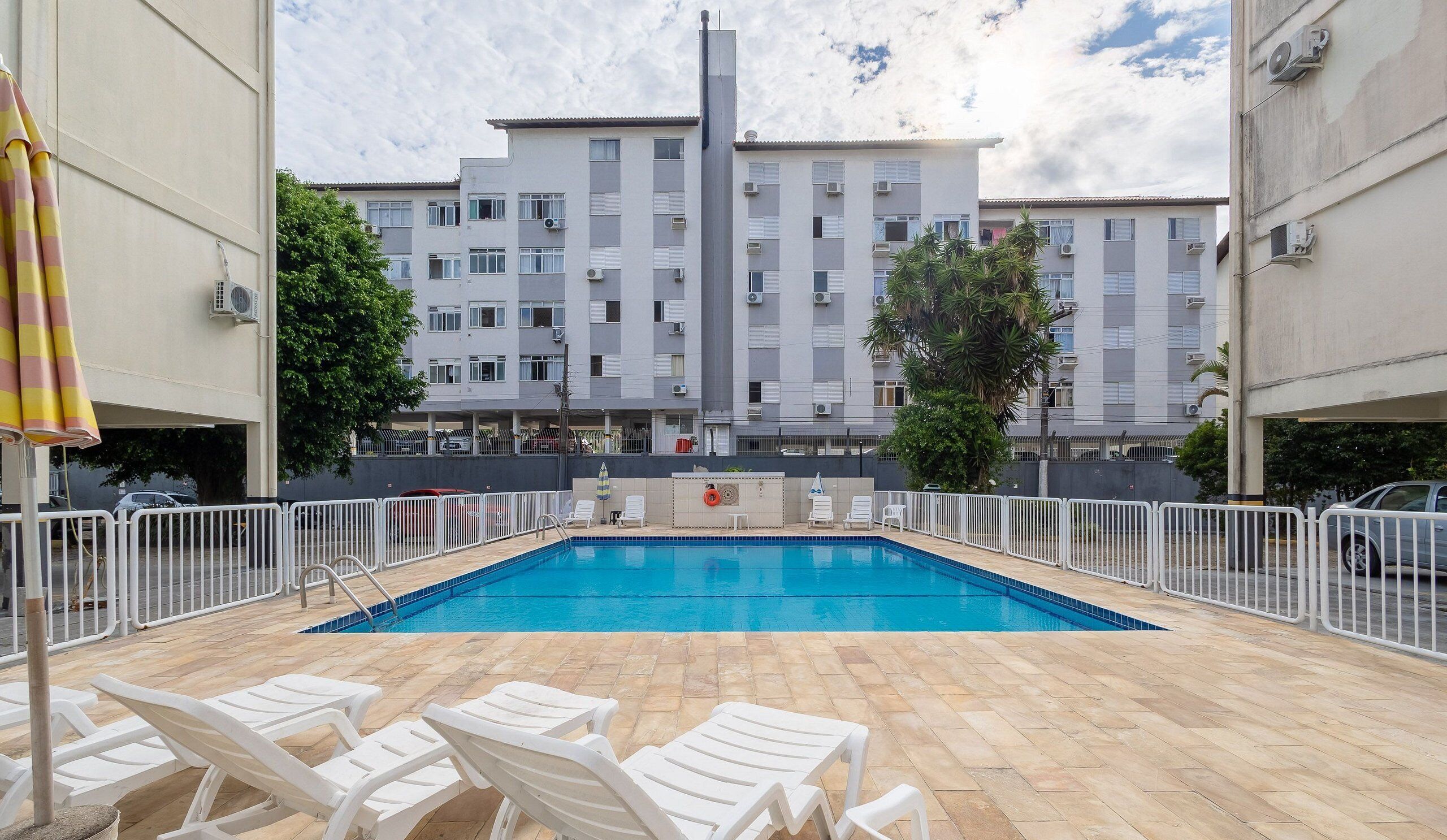 A swimming pool with white lounge chairs, surrounded by a white fence, and a building with multiple balconies in the background.
