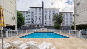 A swimming pool with white lounge chairs, surrounded by a white fence, and a building with multiple balconies in the background.