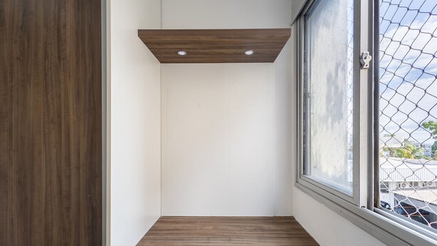 A corner room with a wooden floor, a white wall, and a window with a chain-link fence outside.