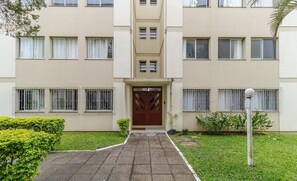 Um edifício de vários andares com entrada central, entrada por portão e gramado bem cuidado.