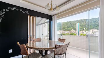 A modern dining area with a glass table and wicker chairs, large windows with a city view, and a black accent wall with a leaf design.