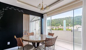 Un espace de repas moderne avec une table en verre et des chaises en osier, de grandes fenêtres offrant une vue sur la ville, et un mur d’accent noir orné d’un motif de feuilles.