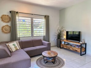 Een woonkamer met een bank, een salontafel, een televisie op een houten dressoir en een groot raam met gordijnen.