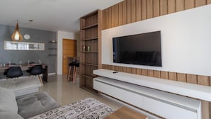 A modern living room with a large flat-screen TV mounted on a wooden wall, a grey sofa, a patterned ottoman, and a dining area with a wooden table and chairs. - Comfort Close to Campeche Beach! - Ddl0206 (Florianópolis)
