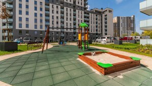 Een speeltuin met schommels en een glijbaan, omgeven door een groene, rubberen ondergrond, voor een hoog appartementencomplex.