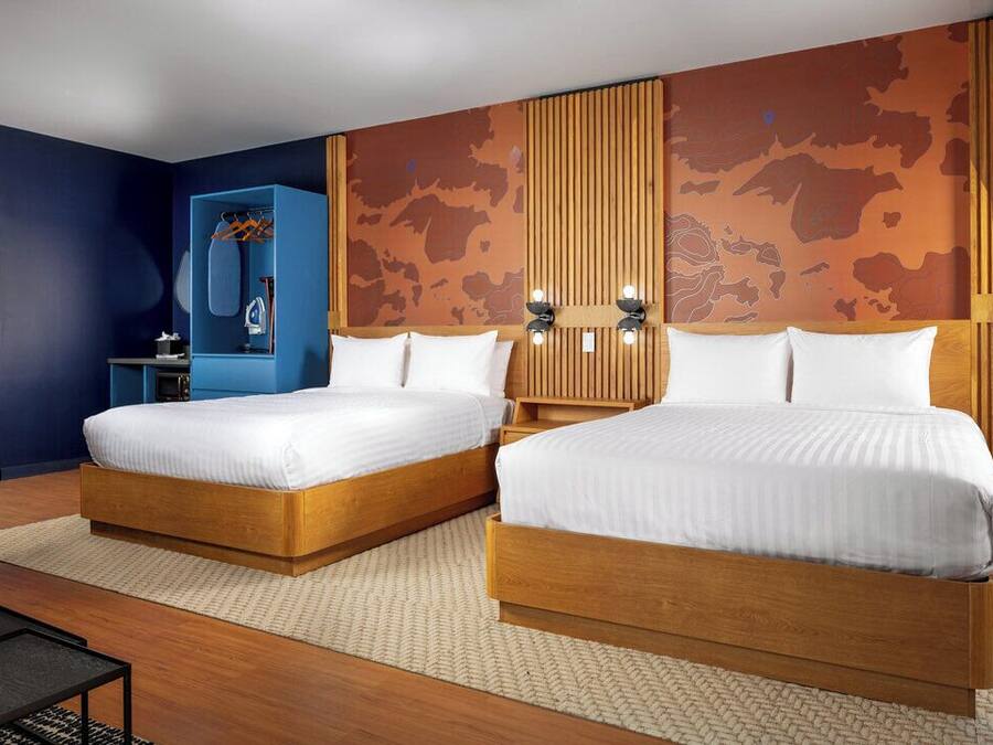Quarto de hotel com duas camas de madeira, um guarda-roupa azul, cabeceira de madeira com luzes e um mapa de parede grande.