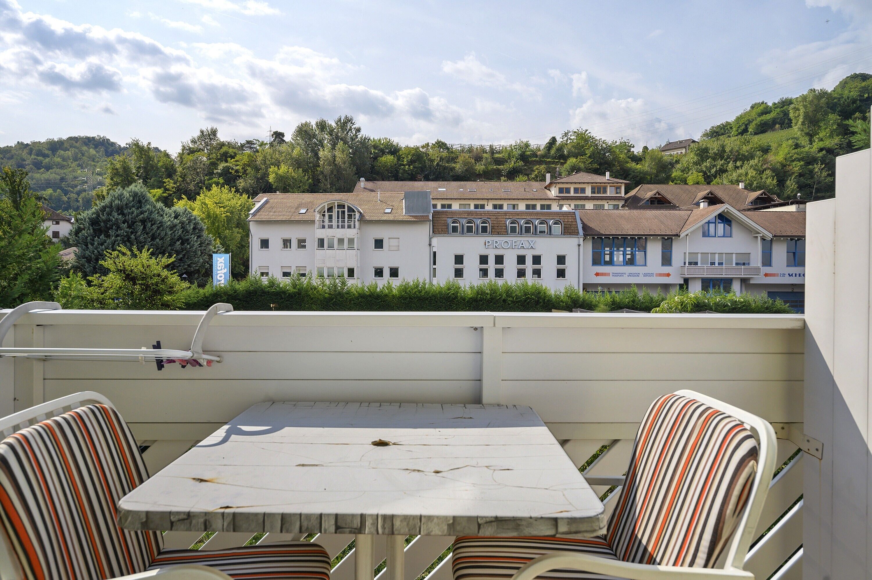 En balkon med et bord og stribede stole, med udsigt over en række bygninger med et skilt der siger 'PROFAX'.