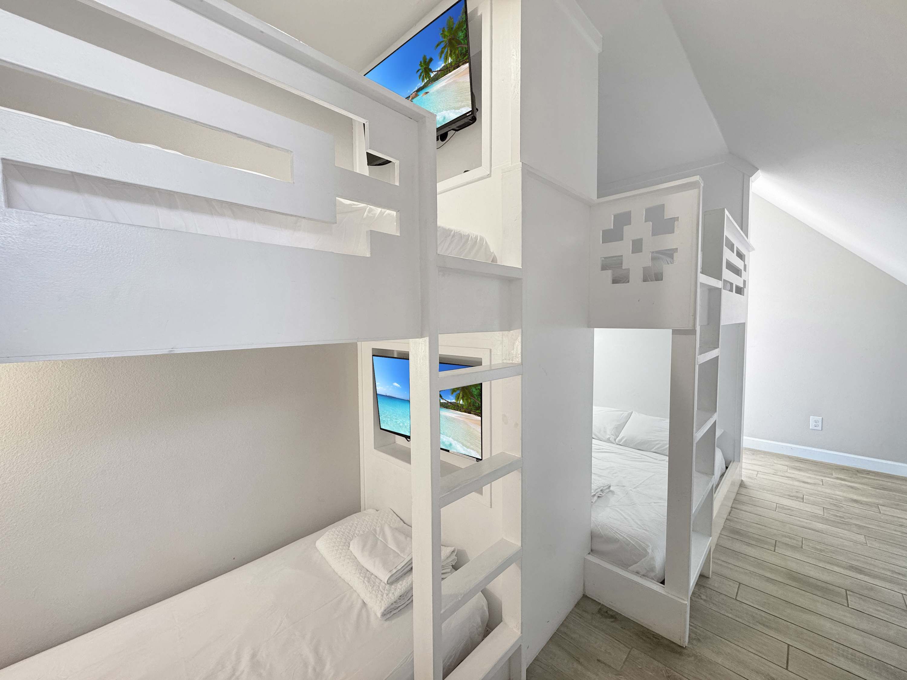 A bunk bed room with a TV mounted above it, a wooden floor, and a view of a beach through a window.