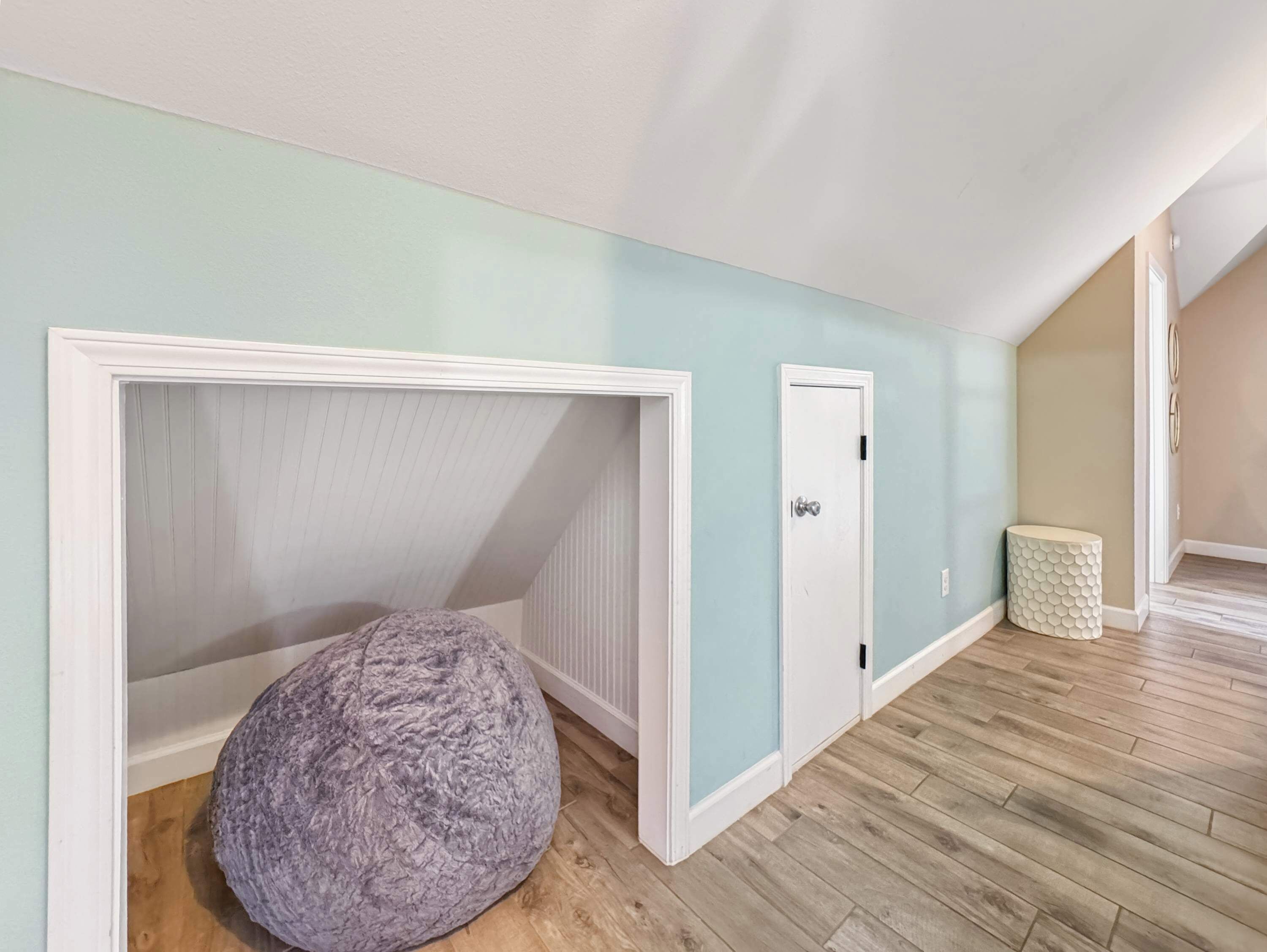 A room with a sloped ceiling, a large grey bean bag chair, a white door, and a textured white side table.