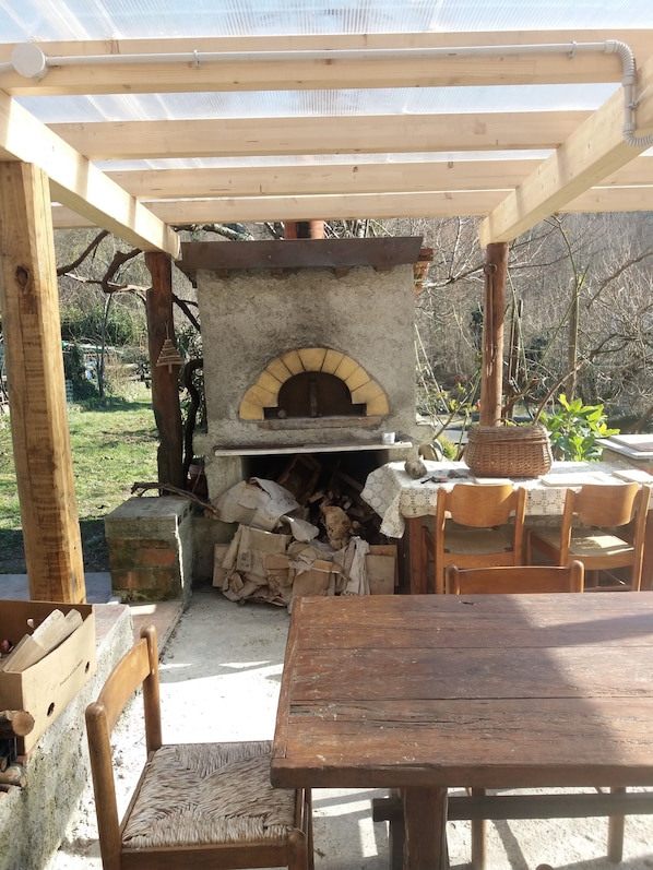 Une pergola en bois installée à l’extérieur, avec un four en briques en dessous, entourée d’une table et de chaises.