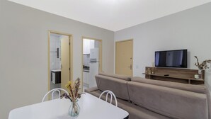 Un salon avec un canapé, un téléviseur et une table à manger avec des chaises. On peut apercevoir la cuisine à travers une porte ouverte.