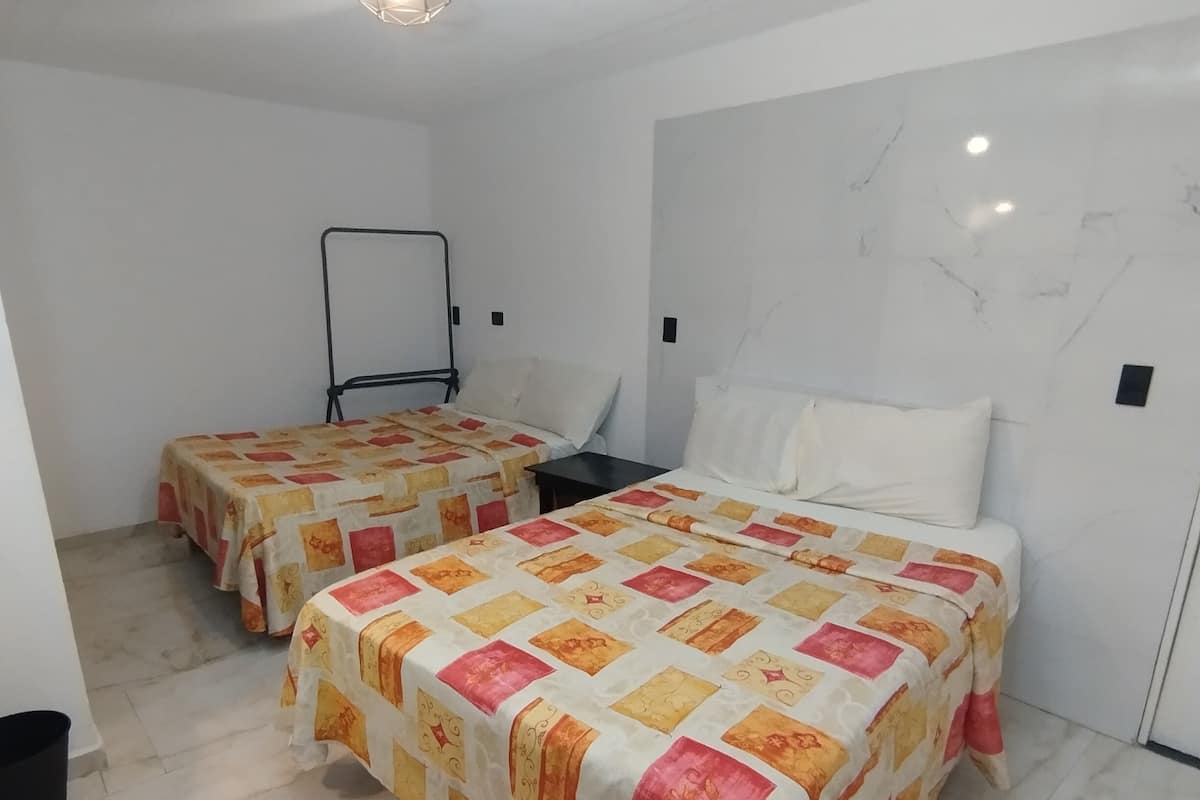 A hotel room with two beds, each covered with a colorful quilt, a black metal bed frame, a white wall with a marble-like texture, and a hanging light fixture.