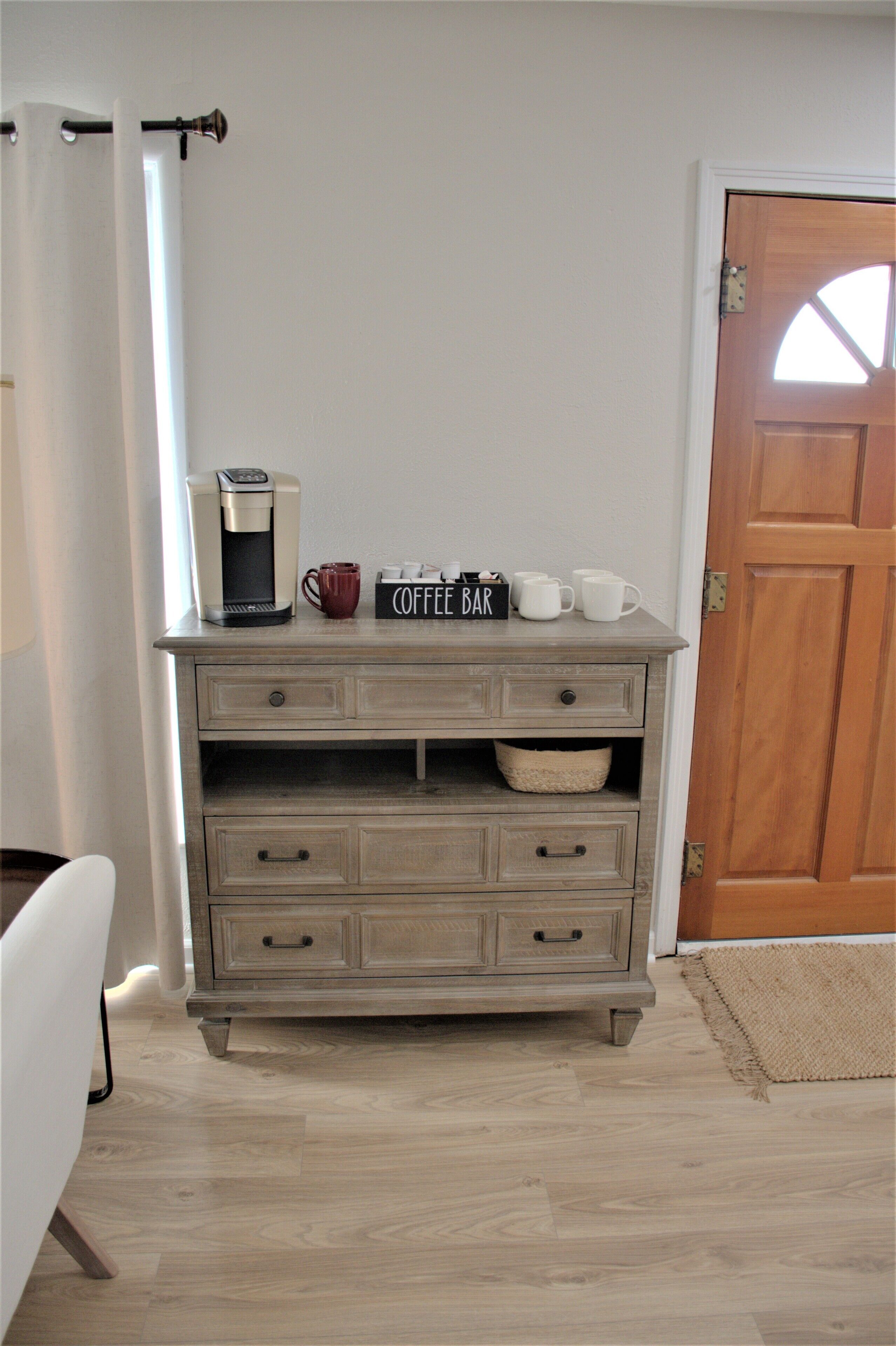 Een houten koffiebar met een koffiezetapparaat, mokken en een bord waarop 'Coffee Bar' staat, in een ruimte met een houten deur en een witte muur.