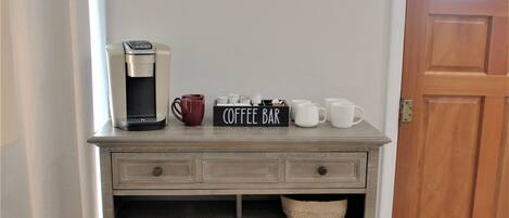 Een houten koffiebar met een koffiezetapparaat, mokken en een bord waarop 'Coffee Bar' staat, in een ruimte met een houten deur en een witte muur.