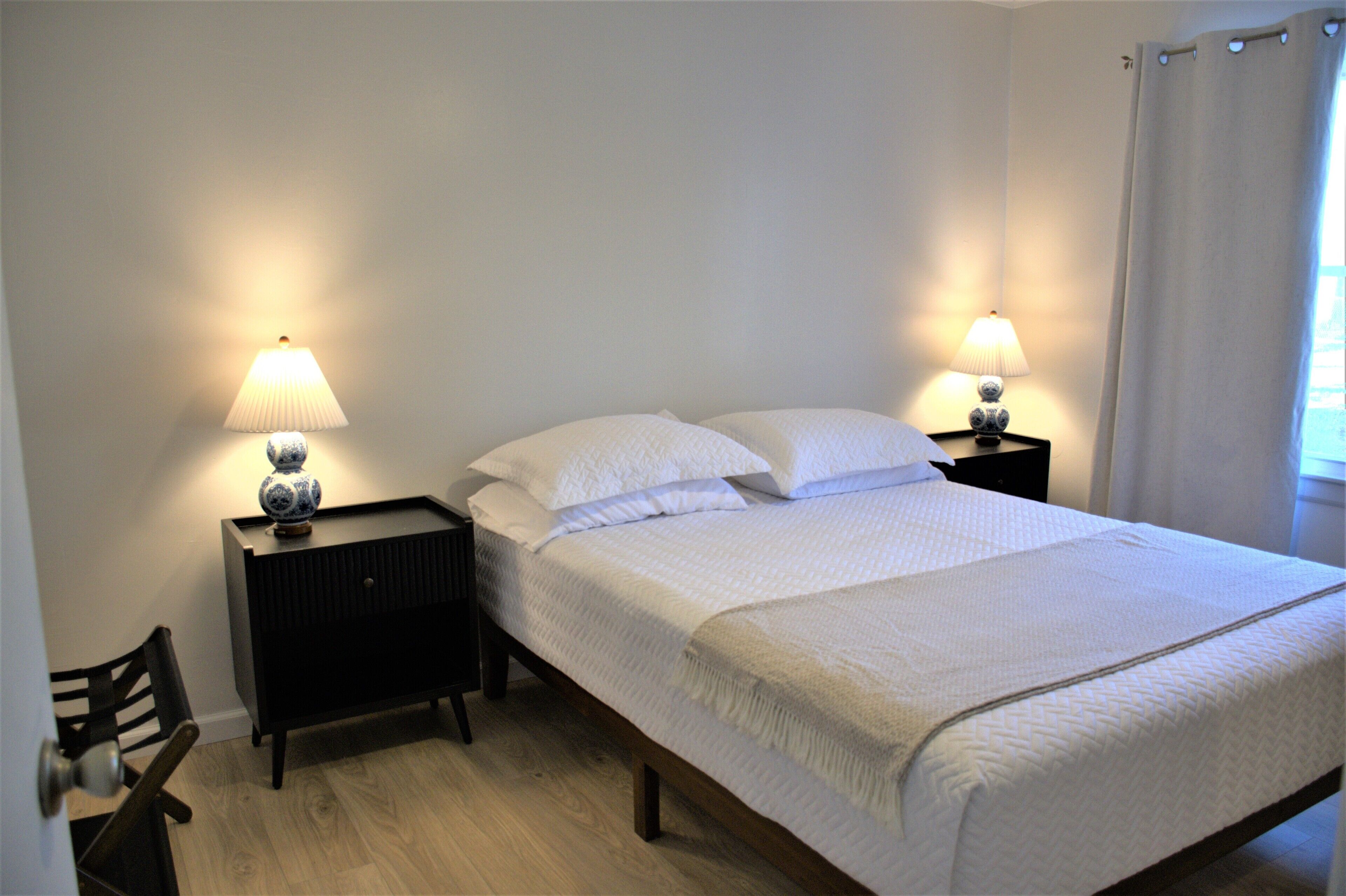 A neatly made bed with white bedding, two bedside tables with lamps, a wooden chair, and a window with curtains.