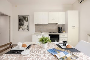Une cuisine moderne avec des meubles blancs, une cuisinière à gaz et une machine à café. On y trouve une table à manger avec une plante, un croissant et un magazine.