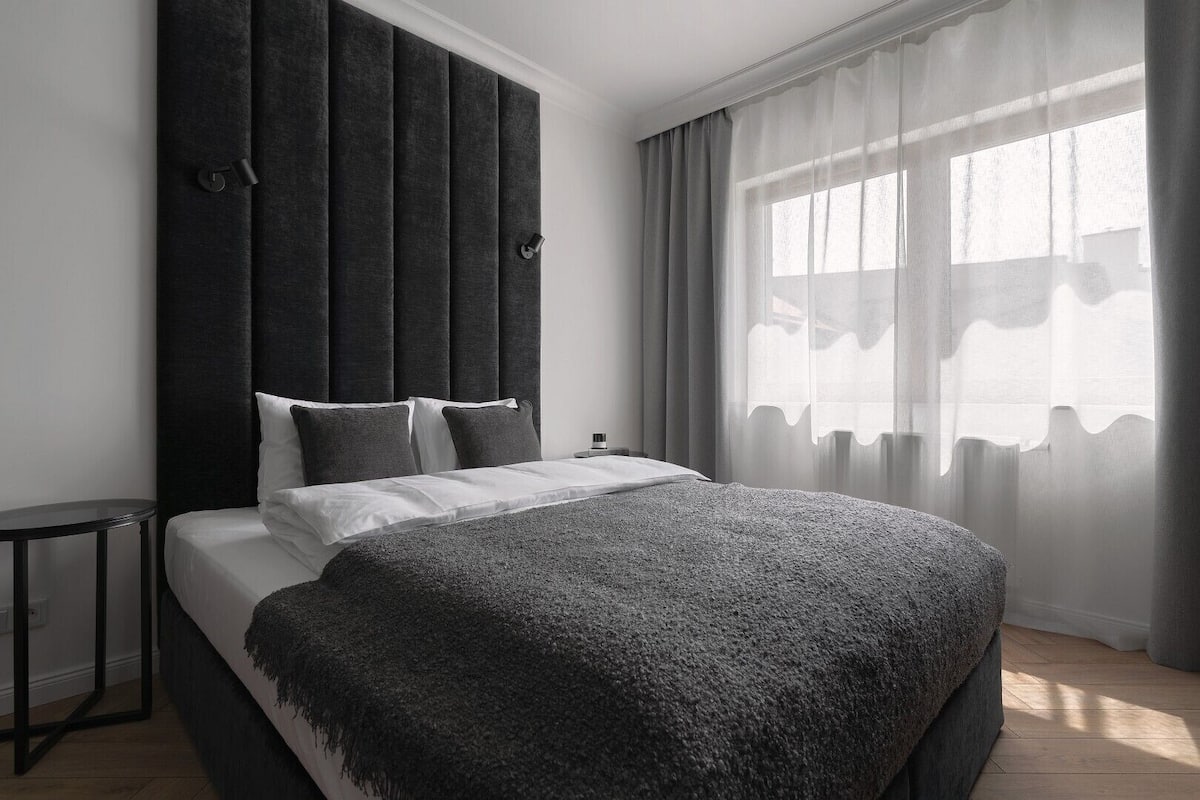 A modern bedroom with a large bed, a dark headboard, and sheer curtains allowing natural light.