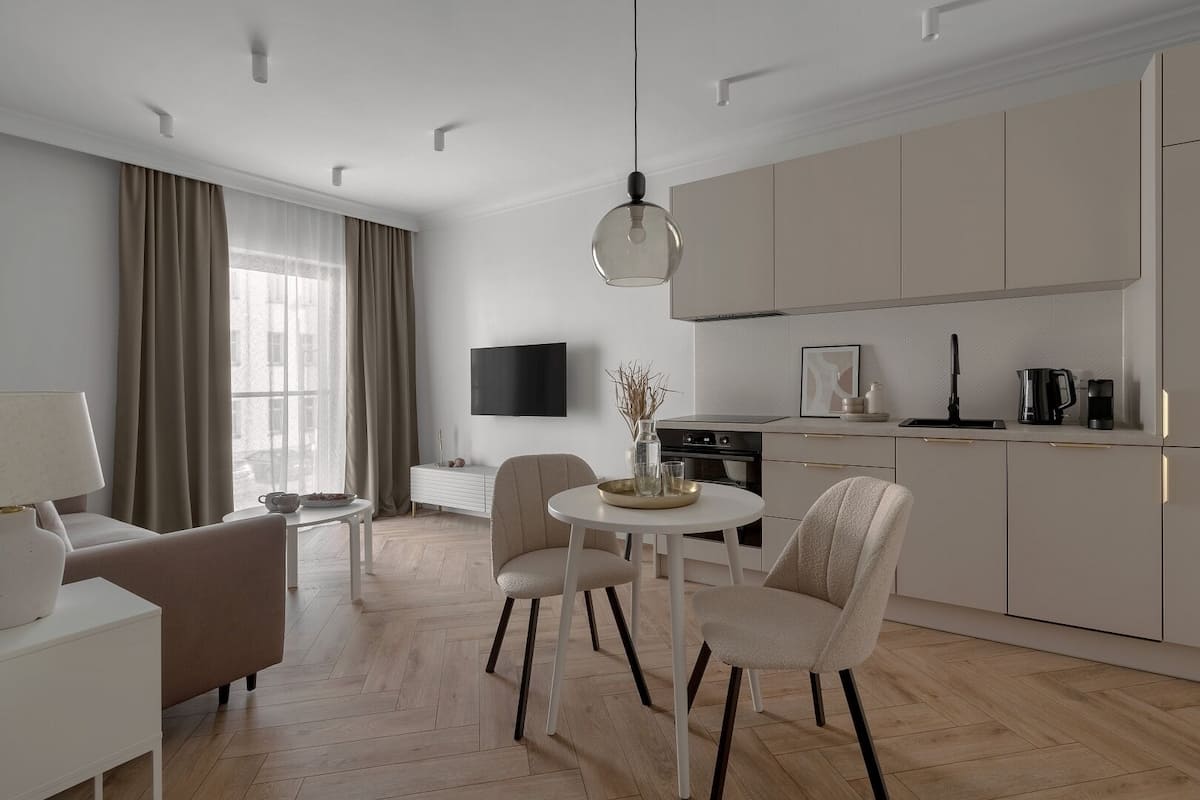 A modern living room with a sofa, a dining table with chairs, a TV mounted on the wall, and a kitchen area with cabinets and appliances.
