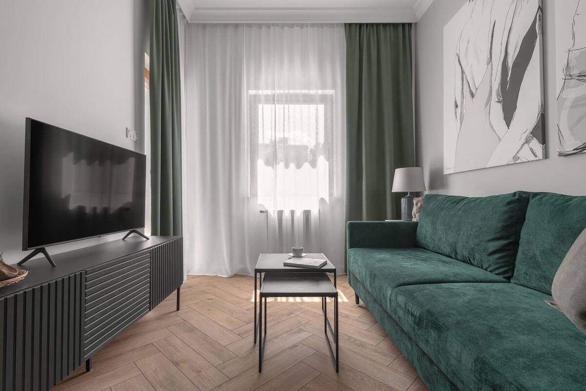 A modern living room with a flat-screen TV on a dark console, a green sofa, a small table with a lamp, and a window with sheer curtains.