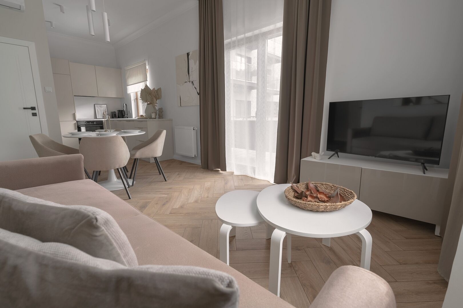 A modern living room with a sofa, two white tables, a TV, and a kitchen in the background.