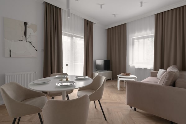 A modern living room with a round dining table, beige sofas, and a flat-screen TV. - Apartio Rooms Gliwice (Gliwice)