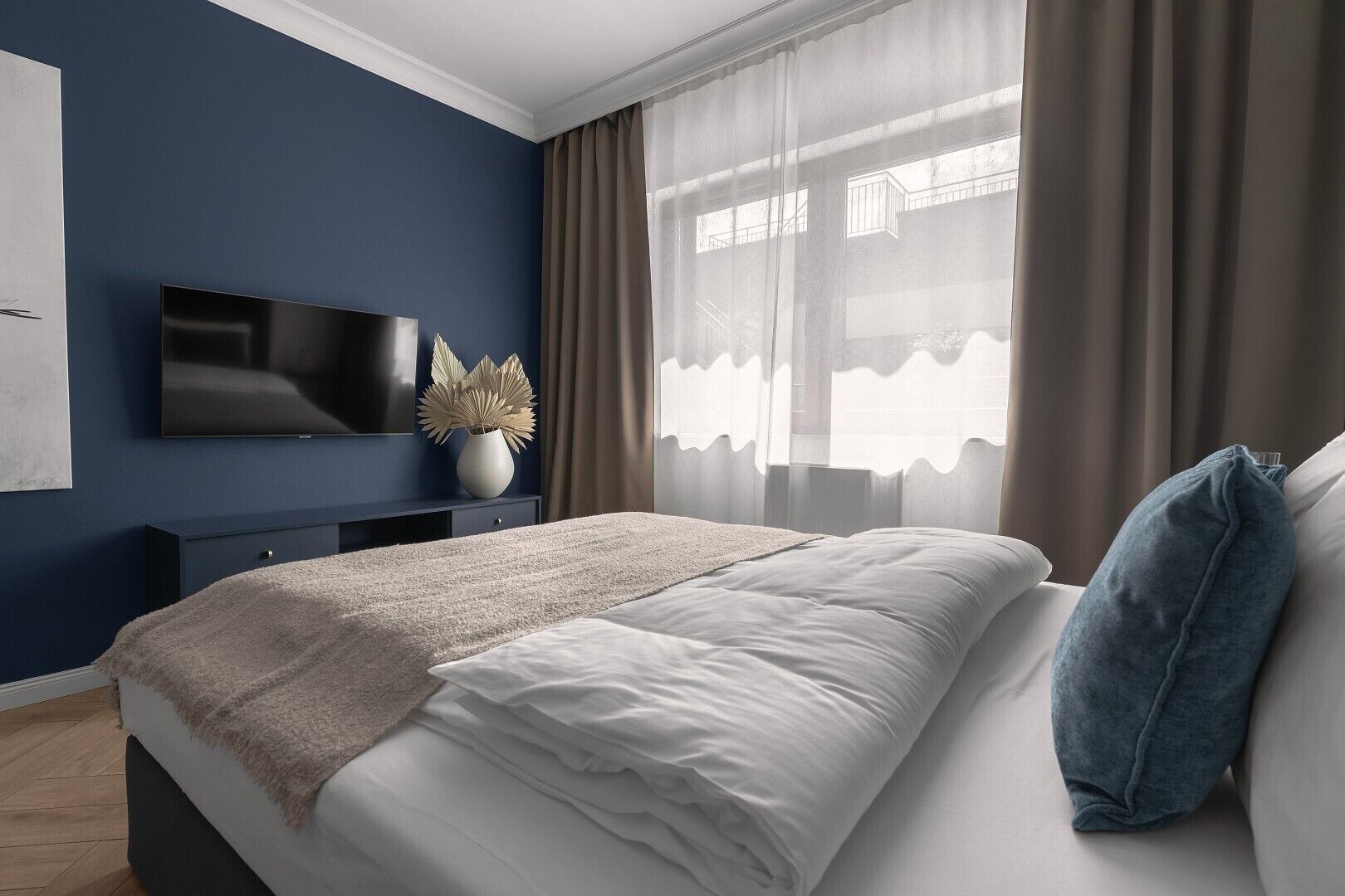 A modern bedroom with a large bed, a flat-screen TV, a vase with dried plants, and sheer curtains.