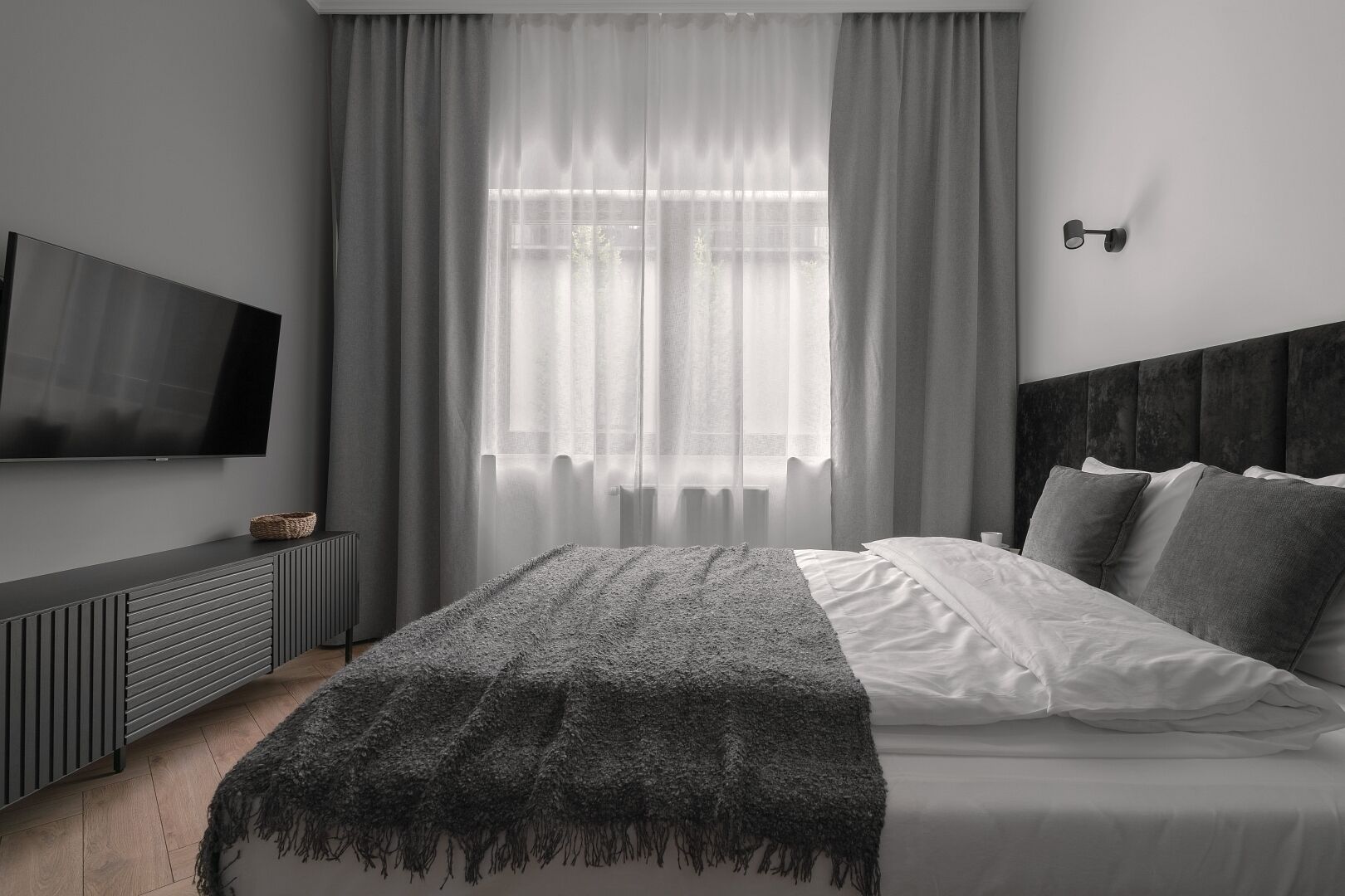 A modern bedroom with a large bed, a TV mounted on the wall, and a window with sheer curtains.