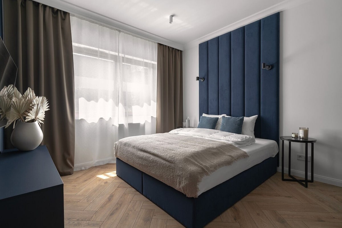 A modern bedroom with a large bed, a bedside table, a nightstand, and a window with sheer curtains.