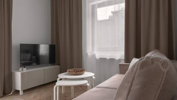 Una sala de estar moderna con un televisor de pantalla plana, una consola blanca, una mesa redonda blanca, un sofá beige y una ventana con cortinas traslúcidas.