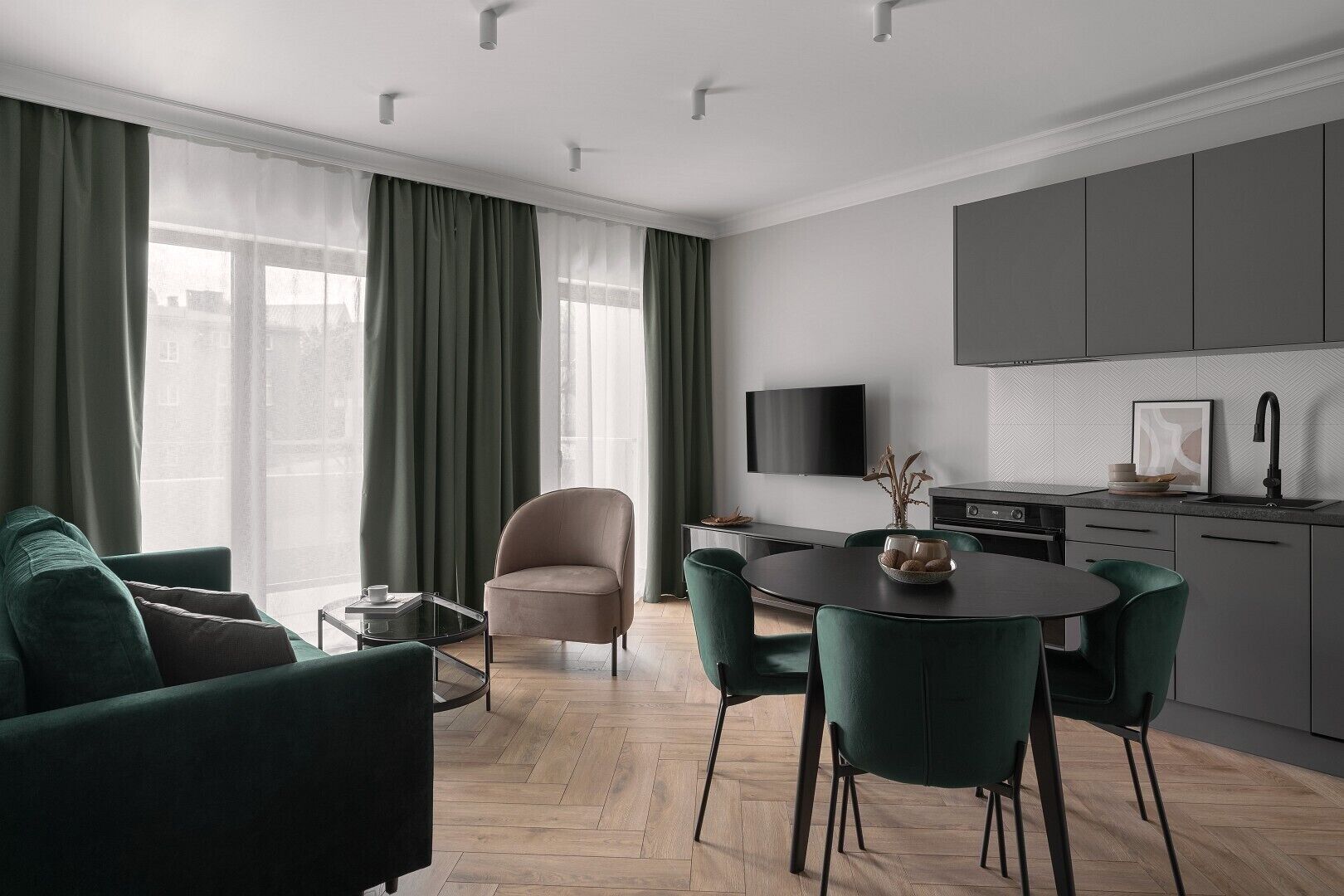 A modern living room with a sofa, armchair, and dining table. There is a kitchen area with a TV and a sink.