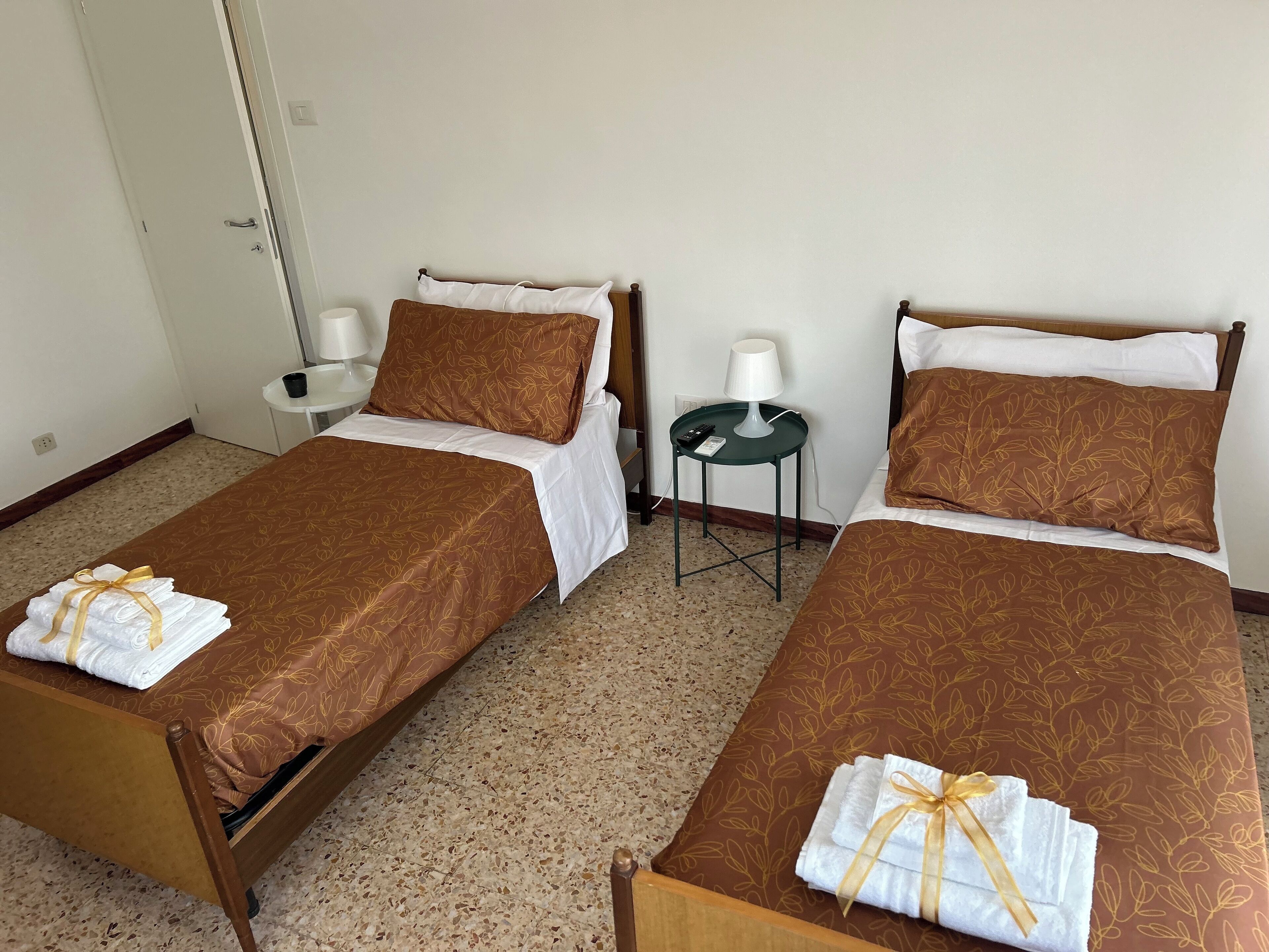 Deux lits simples avec des couvre-lits à motifs floraux marron et des oreillers blancs, une petite table verte avec une lampe, et une chaise blanche.