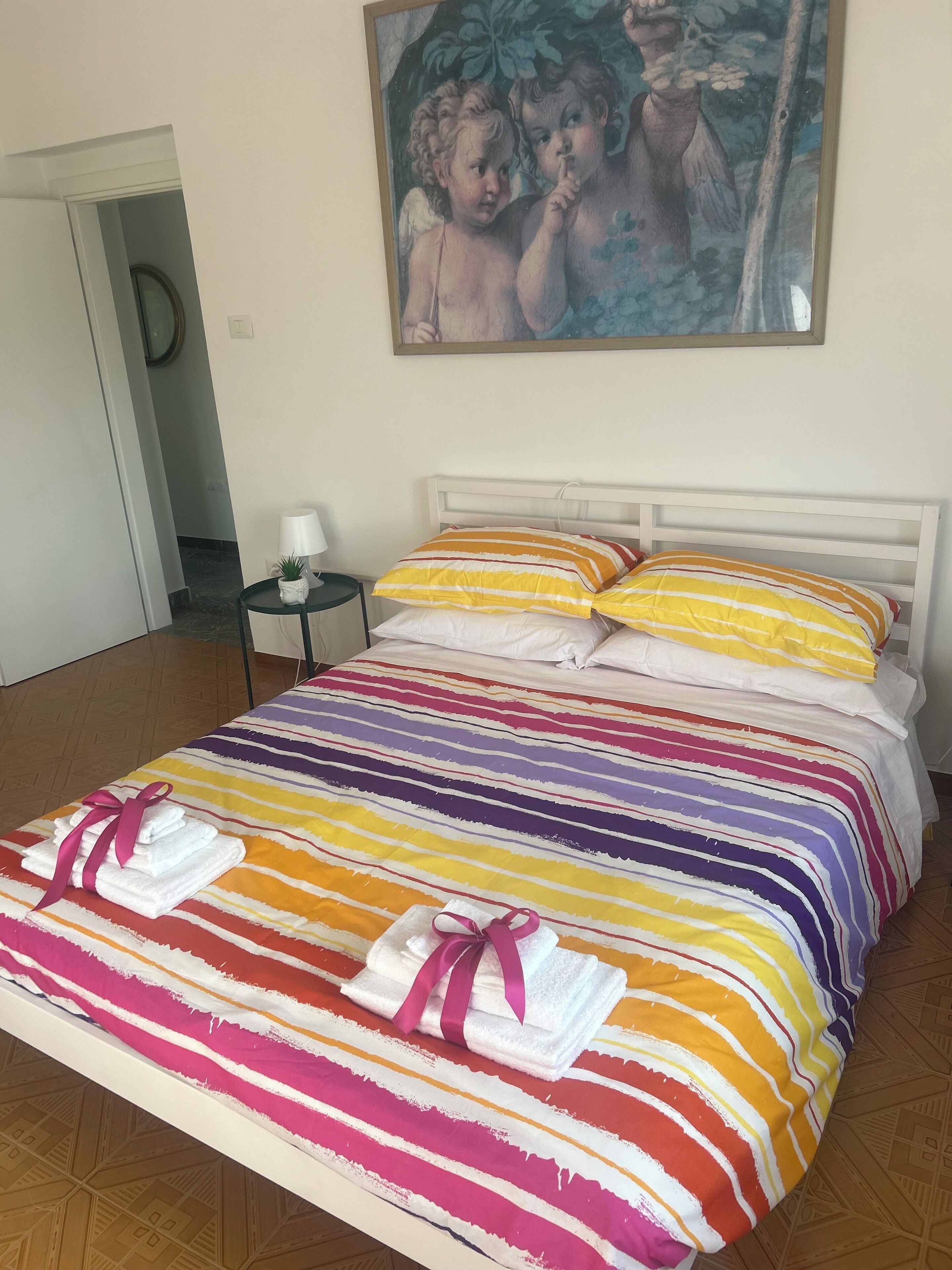 En seng med et farverigt stribet sengetæppe og puder, et lille natbord med en lampe, og et maleri på væggen.