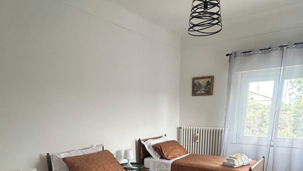Une chambre d’hôtel avec deux lits simples, une petite table ronde, une lampe, un radiateur et une fenêtre avec des rideaux.