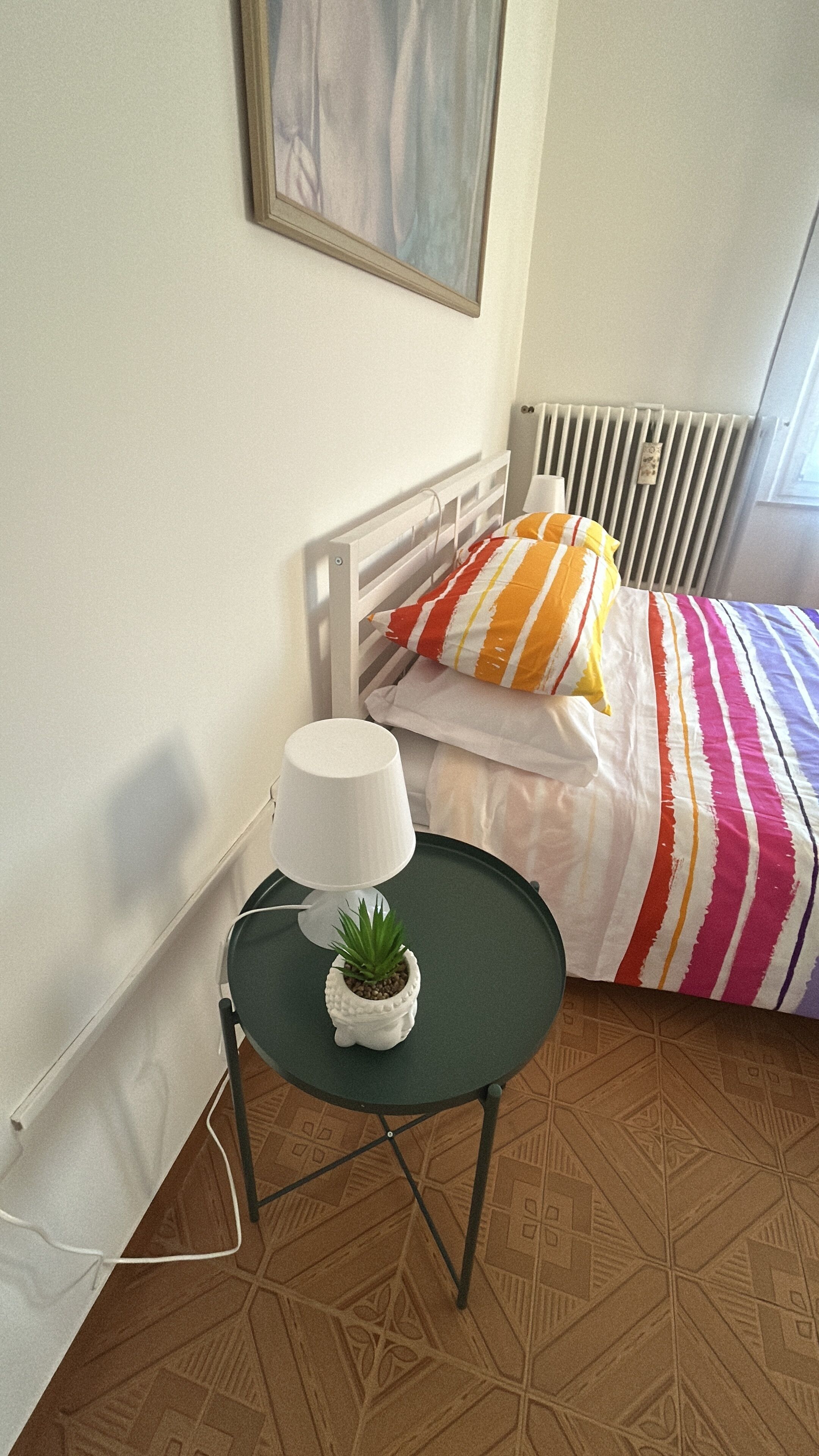 A bedroom with a bed, a bedside table with a lamp and a small plant, a radiator, and a patterned floor.
