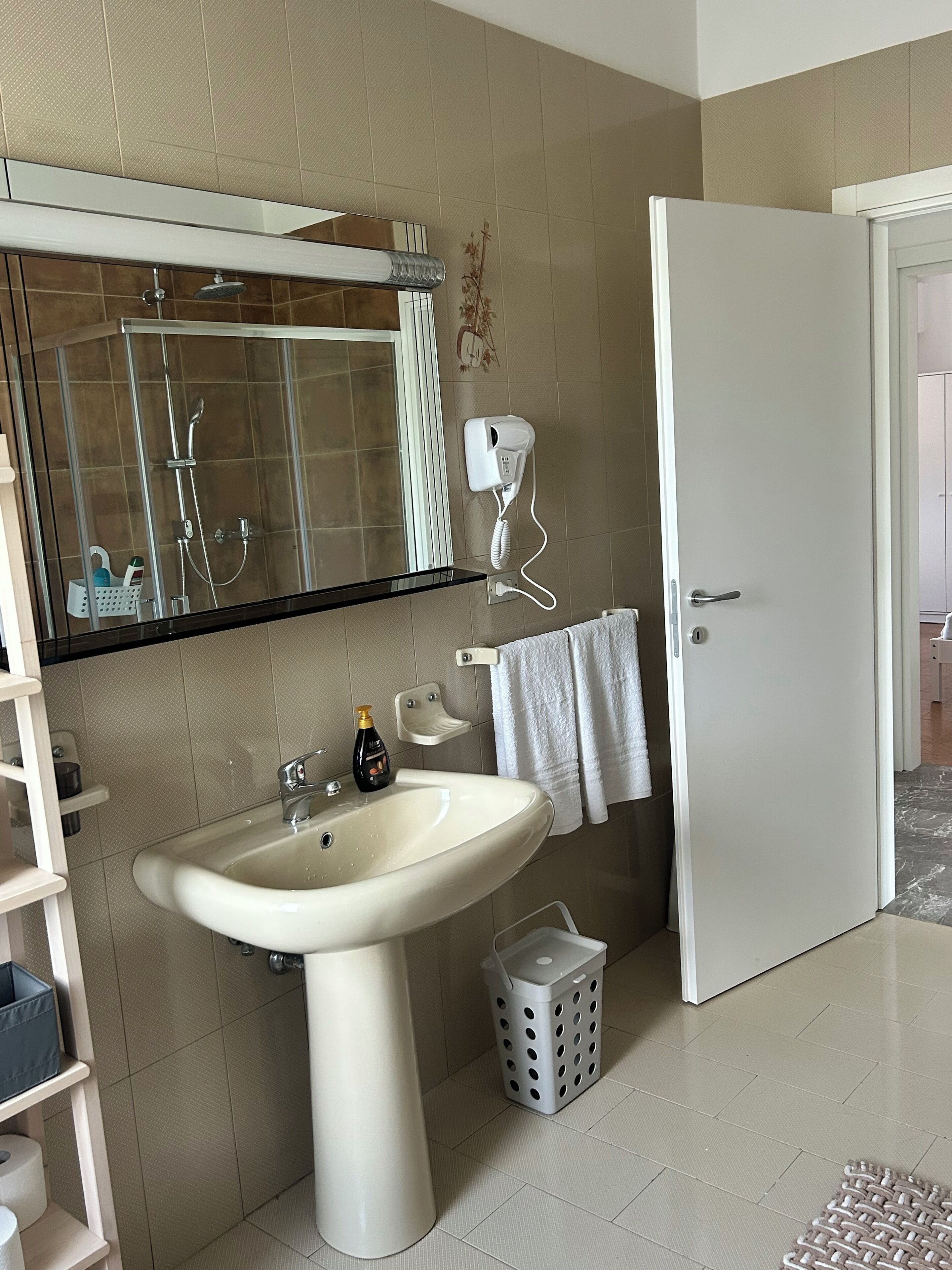 Un bagno con un lavandino bianco, un box doccia in vetro, un asciugacapelli e una porta bianca.