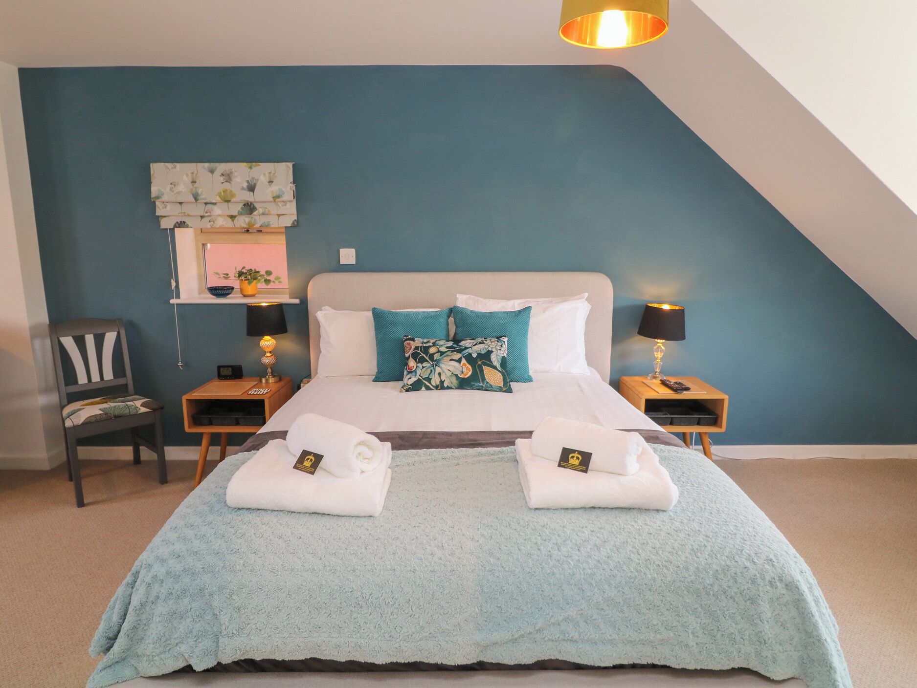 A neatly made bed with two towels and a blanket, a chair, bedside tables with lamps, a wall clock, and a small shelf with a plant.