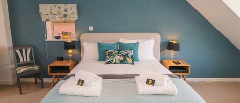 A neatly made bed with two towels and a blanket, a chair, bedside tables with lamps, a wall clock, and a small shelf with a plant.