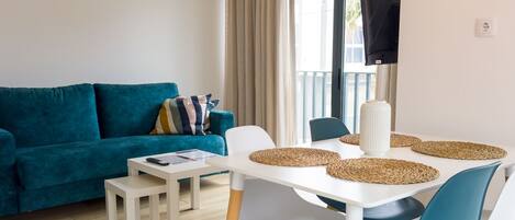 Una sala de estar moderna con un sofá verde azulado, una mesa de comedor blanca con sillas azules y un ventanal con cortinas.