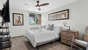 A bedroom with a bed, a ceiling fan, a window with a view of palm trees, a framed picture, a TV on a stand, and a nightstand with a lamp. - Tropical Escape With Pool Tennis Cabo Rojo (Cabo Rojo)