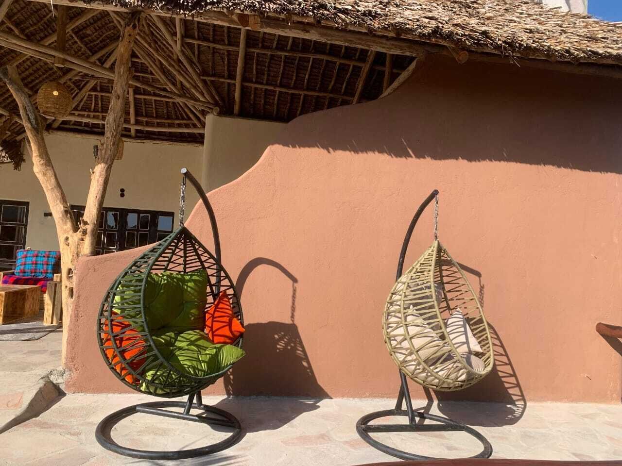 Dos sillas colgantes con cojines frente a una pared, un árbol y un edificio con techo de paja al fondo.
