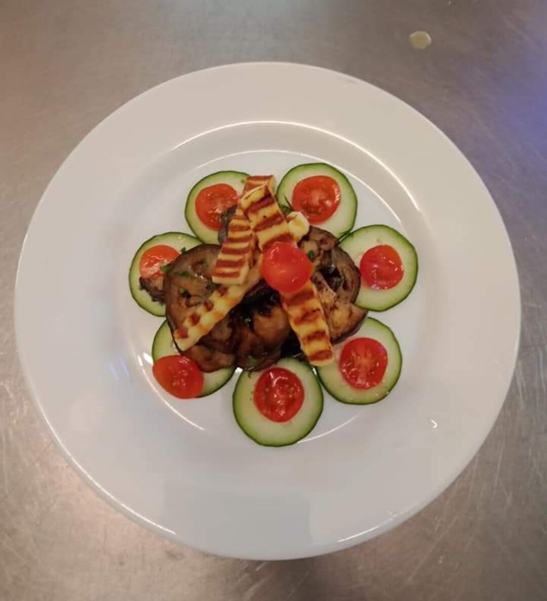 A white plate with grilled chicken, cucumber slices, and cherry tomatoes arranged on a stainless steel surface.