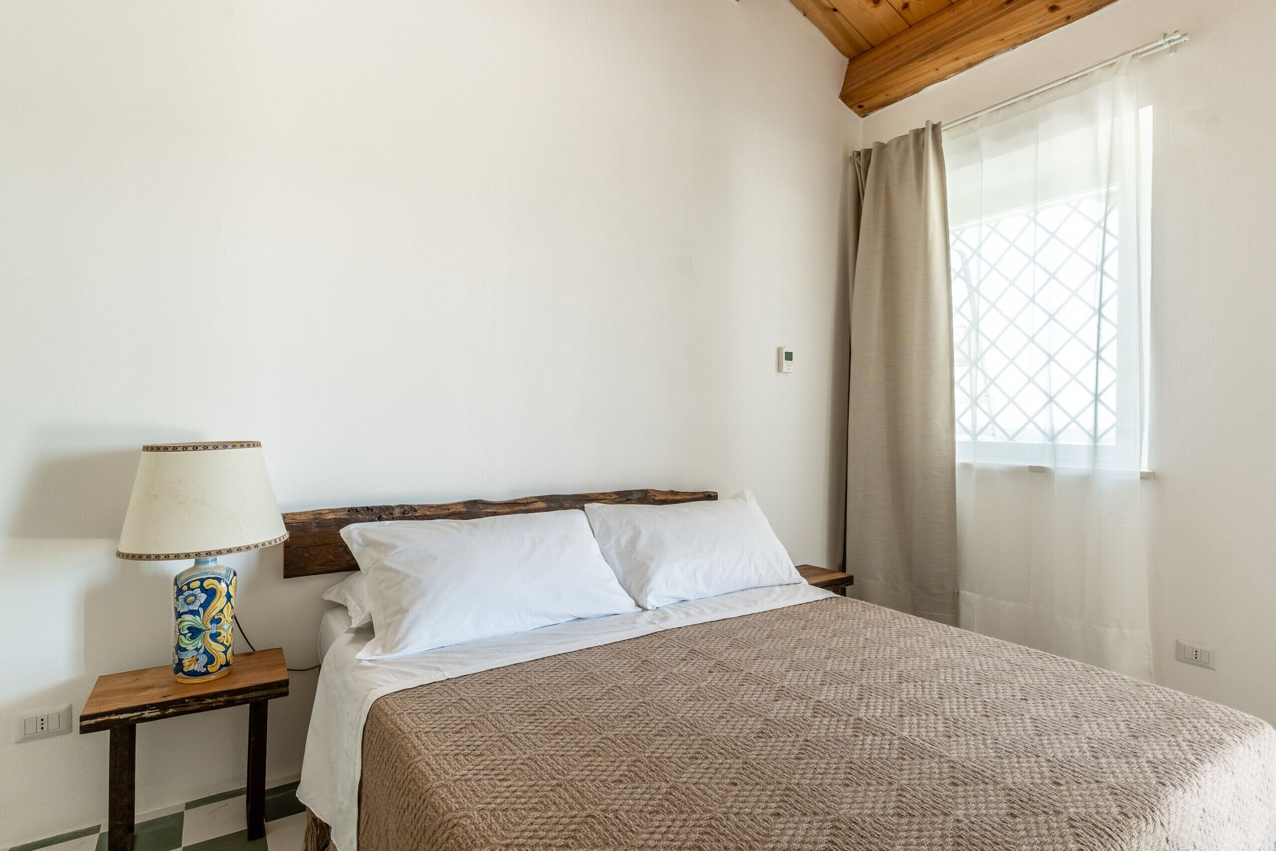 A bedroom with a bed, a nightstand with a lamp, a window with curtains, and a wooden ceiling.