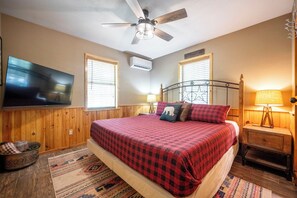 Ein Schlafzimmer mit Bett, Deckenventilator, an der Wand montiertem Fernseher und einem Fenster mit Jalousien.